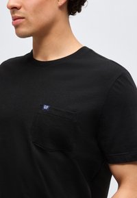 Bărbat purtând un tricou simplu negru cu mâneci scurte și un buzunar pe piept inscripționat cu „GAP” pe un fundal gri simplu.
