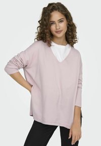 Pull léger rose, ample, avec un décolleté en V et des manches longues, porté par-dessus une chemise blanche. Sa texture est douce et il possède des coutures d'épaule tombantes.