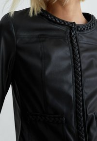 Czarna skórzana kurtka z plecionym kołnierzem i ozdobami na kieszeniach. Strukturalny krój z gładką fakturą i starannym przeszyciem.