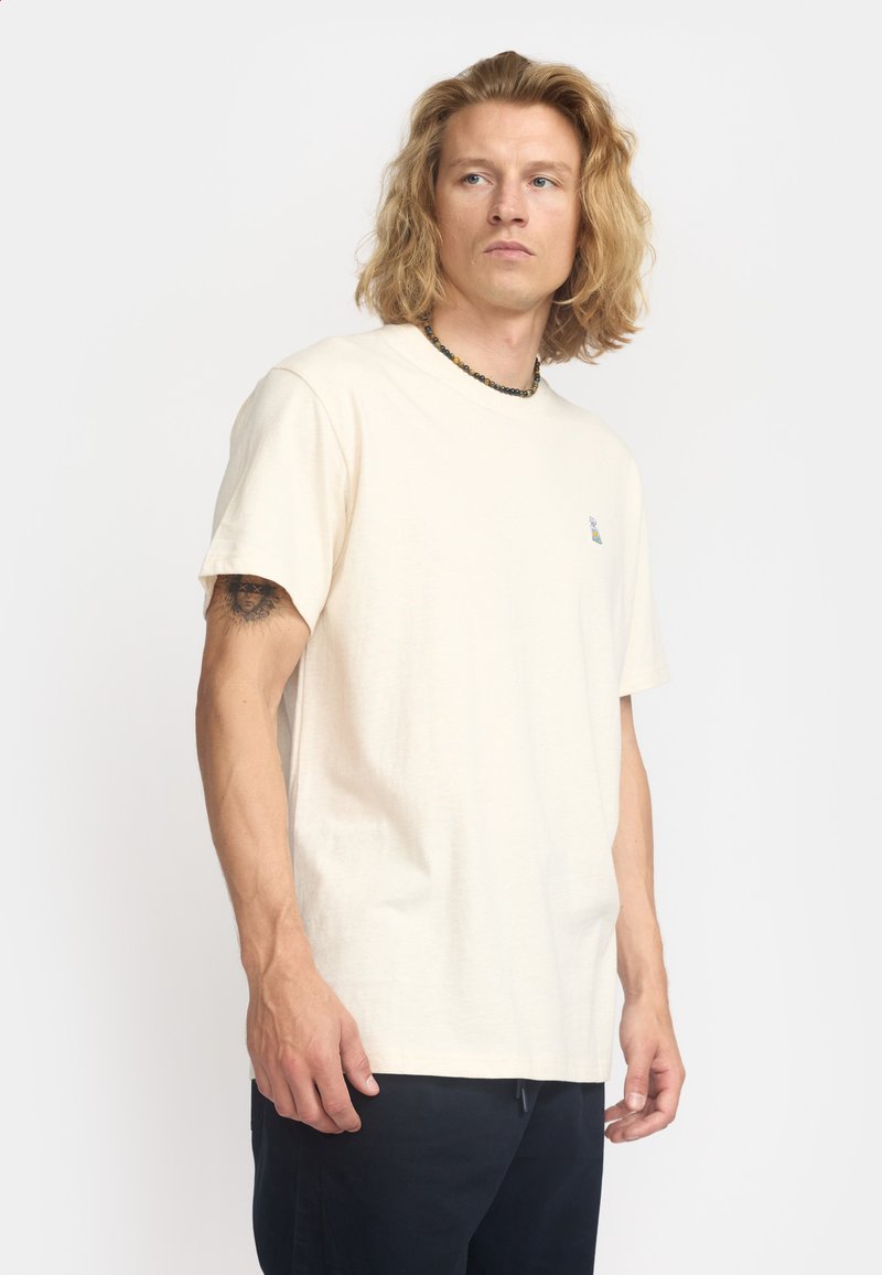 Camiseta de algodón beige con cuello redondo y mangas cortas; presenta un pequeño logotipo bordado colorido en el lado izquierdo del pecho.