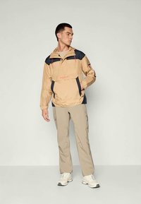 Tan vanntett jakke med marineblå detaljer, frontlomme, justerbar hette og en løs passform, sammen med lys beige bukser og hvite joggesko.