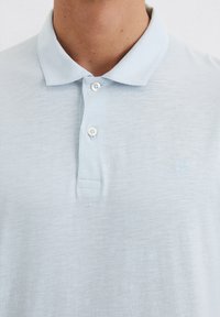 Lichtblauwe polo met een gestructureerde afwerking, voorzien van een kraag, een drieknopen-sluiting en een subtiel geborduurd logo op de borst.