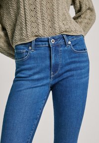 Vaqueros de mezclilla azul ajustados con una textura suave, diseño de cinco bolsillos y costuras en contraste. Accesorios de metal plateado adornan la cinturilla.