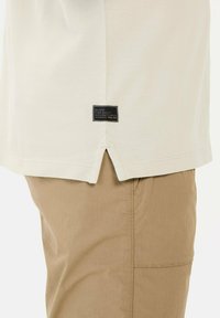 Cremefarvet stof t-shirt med en sideslids og et sort mærke med hvid og orange tekst syet nær bunden. Båret med beige bukser.