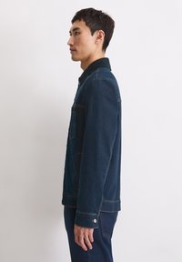 Donkerblauwe denimjack met een klassieke kraag, een voorklepzak en oranje stiksels; gedragen over bijpassende broek, weergegeven in profielweergave.