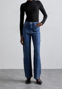 Henkilö, joka on pukeutunut mustaan, pitkähiharaiseen, kimalteilla varustettuun toppiin, sinisiin korkeavyötäröisiin suoriin farkkuihin, joissa on kimallusta, sekä mustiin, kärjeltä�än teräviin korkokenkiin, joissa on soljet.