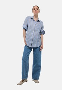 Chemise boutonnée rayée bleue et blanche avec manches retroussées, associée à un jean large bleu clair et des ballerines beiges.