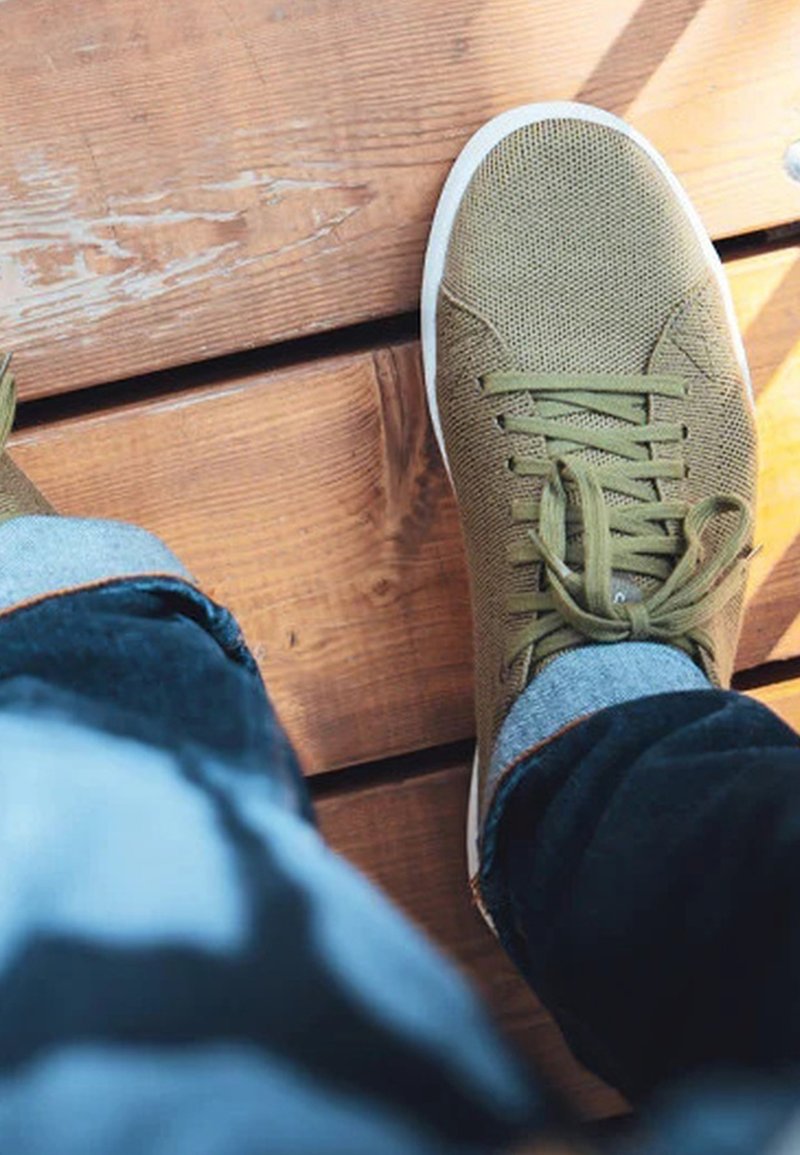 Pie derecho con una zapatilla verde y jeans azules remangados sobre una plataforma de madera, con patrones de luz y sombra.