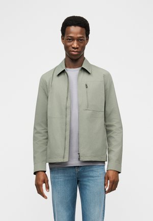 Muž ve světle šedé bundě na zip přes šedé tričko a modré džíny, stojící před jednobarevným bílým pozadím a čelící dopředu.