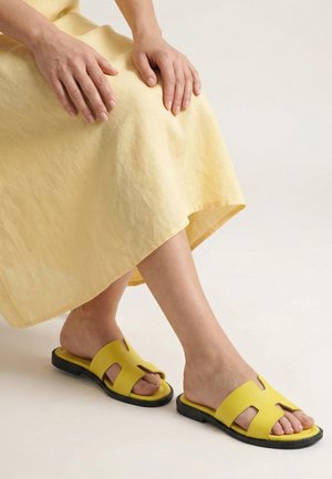 Person trägt ein gelbes Kleid und gelbe Slip-On-Sandalen mit schwarzen Sohlen, sitzt mit Händen auf dem Schoß.