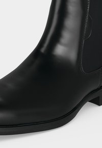 Gros plan d'une botte Chelsea en cuir noir avec un panneau latéral élastique et un talon bas sur fond blanc.