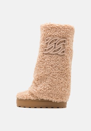 Beige pörröinen nilkkuri, jossa on teksturoitu pinta, korostettuna selkeällä logolla ja tukevalla kumipohjalla, joka tarjoaa pitoa.