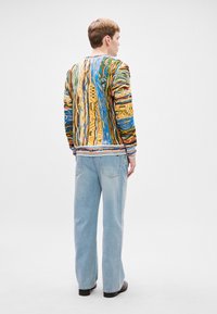 Värikäs kuviollinen neule, jossa on sinisiä, keltaisia, vihreitä ja punaisia grafiikoita. Yhdistettyä vaaleansinisiin leveälahkeisiin farkkuihin ja mustiin kenkiin.