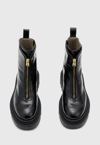 Botas de cuero negro hasta el tobillo con un zipper dorado, puntera redondeada y suela gruesa negra. Cuenta con una lengüeta en la parte posterior para facilitar su uso.