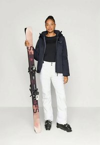 Sprzęt narciarski: osoba w granatowej kurtce, czarnej bluzce, białych spodniach, trzymająca różowe i czarne narty z wiązaniami, na jasnym tle.