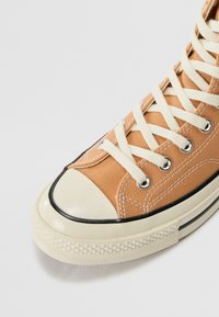 Zbliżenie beżowego trampka z białymi sznurówkami, białą gumową czubką i teksturowaną beżową podeszwą na jasnym tle.