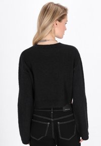 Schwarzer, kurz geschnittener Pullover mit weicher Textur, weiten Ärmeln und geripptem Saum, kombiniert mit schwarzen Jeans mit weißen Stitching-Details.