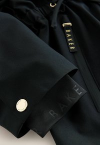 Detail čierneho rukáva bundy so zlatým gombíkom popísaným „Baker by Ted Baker“ a čiernym ťaháčikom zipsu s nápisom „BAKER“.