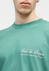 Κοντινό πλάνο ενός ατόμου που φοράει μπλουζάκι teal με λευκό κείμενο που γράφει «Υπό εξέλιξη» και «ΤΟΠΙΚΟ • ΠΑΓΚΟΣΜΙΟ • ΔΙΕΘΝΕΣ • ΠΑΝΚΟΣΜΙΟ».