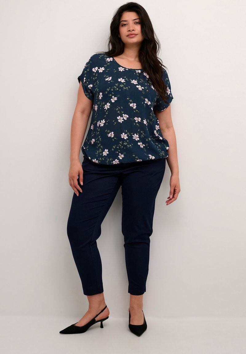 Blouse florale navy à manches courtes, coupe ample ; associée à un pantalon navy slim. Des escarpins à talons aiguilles noirs complètent la tenue.