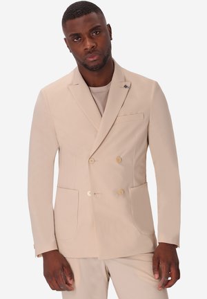 Uomo che indossa una giacca doppiopetto beige e pantaloni abbinati, in piedi davanti a uno sfondo chiaro e uniforme.