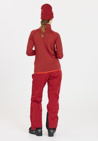 Rotes Thermoshirt mit einem Viertelreißverschluss und gerippter Textur, kombiniert mit roten Schneehosen, die einen kontrastierenden schwarzen Akzent am Saum aufweisen.