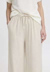 Pantalons larges beige en tissu léger, avec une taille élastique, un cordon de serrage et des poches latérales. Texture douce et design épuré.