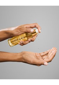  held met een heldere glazen fles met gouden accenten die olie bevat, wordt uitgepompt in een hand. Lichtreflecties en druppels zijn zichtbaar.