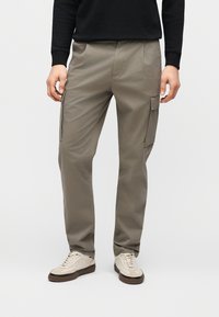 Άνδρας που φοράει μπεζ παντελόνι φόρμας με πλαϊνές τσέπες, μαύρο μακρυμάνικο μπλουζάκι και μπεζ αθλητικά παπούτσια, στέκεται μπροστά σε απλό φόντο.