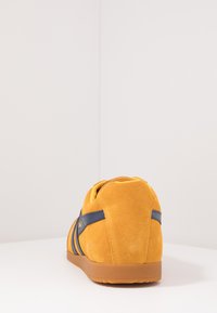 Sneaker giallo in suede con strisce laterali blu, superficie testurizzata e tacco arrotondato. Presenta una suola in gomma e un collo imbottito.