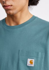 Camiseta de algodón color teal con escote redondo y un bolsillo en el pecho que presenta una etiqueta blanca con un logotipo naranja. Textura suave y diseño casual.