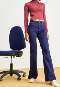 Tuah jeans biru melebar dengan pinggang tinggi dan saku depan, dipadukan dengan atasan lengan panjang marun yang dipotong, berdiri di samping kursi kantor biru.