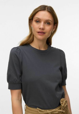 Woman with light brown hair wearing silver hoop earrings, a dark gray puff-sleeve shirt, and beige pants with a fabric flower belt.