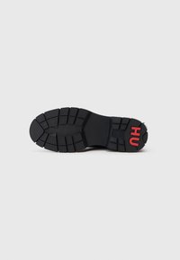 Suela de zapatilla negra con un patrón de textura antideslizante y ranuras profundas; presenta el logo rojo "HU" en el talón como detalle adicional.