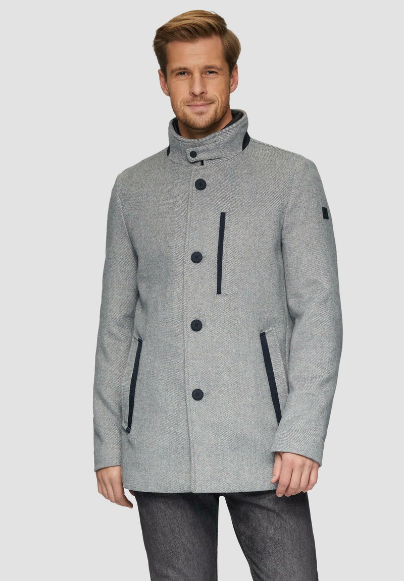 Grijze wolmix jas met hoge kraag, zwarte knopen, twee zijzakken en marineblauwe accenten op de borst, getoond op een model tegen een lichte achtergrond.