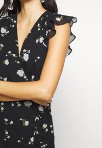 Žena v černých květovaných šatech s volánkovými krátkými rukávy a visícími průhlednými kuličkovými náušnicemi, se zkříženýma rukama před bílým pozadím.