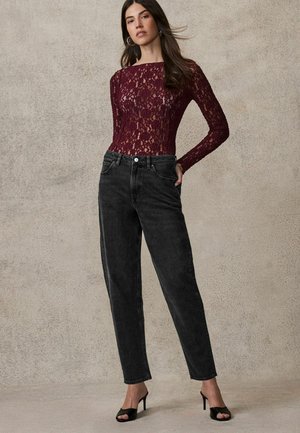 Burgunderfarbener Spitzenbody mit langen Ärmeln, kombiniert mit hoch taillierten schwarzen Jeans mit geradem Bein und schwarzen offenen High Heels, vor einem strukturierten Hintergrund stehend.
