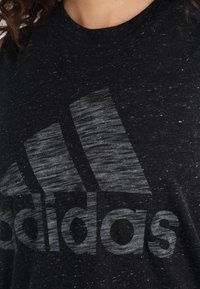 T-shirt noir texturé avec un logo Adidas gris proéminent. Le tissu présente un motif tacheté, avec un col rond.