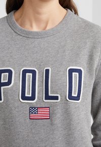 Grå sweatshirt med "POL"-bokstäver i marinblått, inramat i vitt, med en broderad amerikansk flagga nedanför texten. Lättviktig, rundad halsdesign.