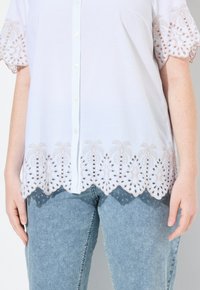 Weißes Hemd mit Knöpfen, das über einen detaillierten beige gestickten Saum mit ausgeschnittenen Designs an Hemdsaum und Ärmeln verfügt, kombiniert mit hellblauen Jeans.