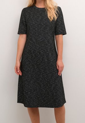 Woman wearing knee-length, short-sleeve black dress with white speckled pattern, standing against plain light background.