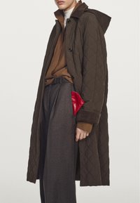 Kviltet brun lang frakke med hætte, med sorte knapper og en rød lommeaccent. Bæres over en brun sweater og grå bukser.