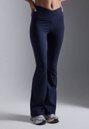 Navy blue flared pants with a wide crossover waistband, made of stretchy fabric. Worn with silver athletic shoes. Smooth texture.