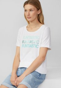 T-shirt en coton blanc à manches courtes, avec un motif de texte métallique turquoise répétitif indiquant "FUNTASTIC".