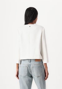 Weiße, kurze Jacke mit Dreiviertelärmeln, strukturiertem Stoff und einem Knopfdetail auf der Rückseite, kombiniert mit hellen Denim-Jeans.
