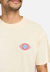 Camiseta de algodón color crema con un gráfico de ojo estilizado en rosa y azul con acentos de líneas radiales, mangas cortas y cuello redondo.