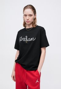 T-shirt in cotone nero con logo "Jordan" in strass bianchi e dettagli dorati. Abbinato a pantaloni tuta rossi con logo Jumpman bianco.