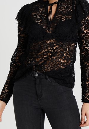Mujer que lleva una blusa negra de encaje transparente con mangas largas, cuello con lazo y vaqueros negros de talle alto.