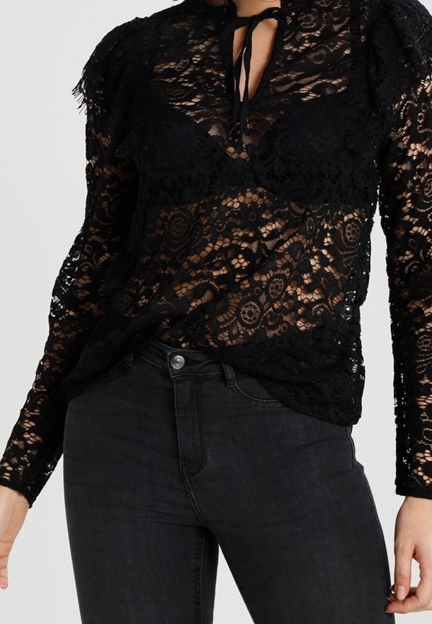 Femme portant un haut long en dentelle noire transparente avec un col à nouer et un jean noir taille haute.
