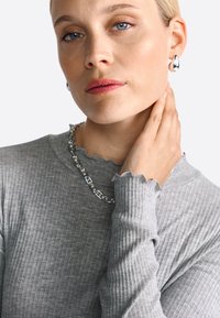 Szürke bordázott, hosszú ujjú felső fodros nyakkivágással, láncnyaklánccal és fényes csepp fülbevalóval párosítva. Sima bőrfelszín, semleges smink.